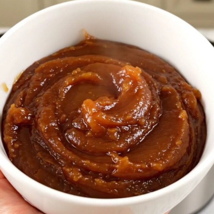 A white bowl filled with glossy, dark brown apple butter, steaming slightly in front of blurred kitchen cabinets.