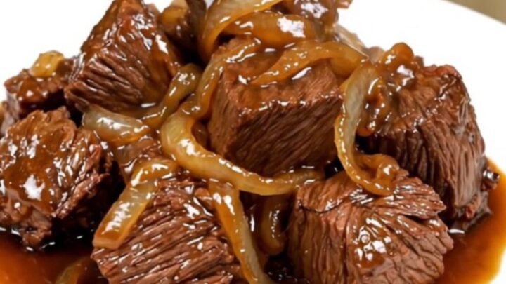 A close-up of tender beef chunks and caramelized onions served on a white plate with a glossy, savory sauce.