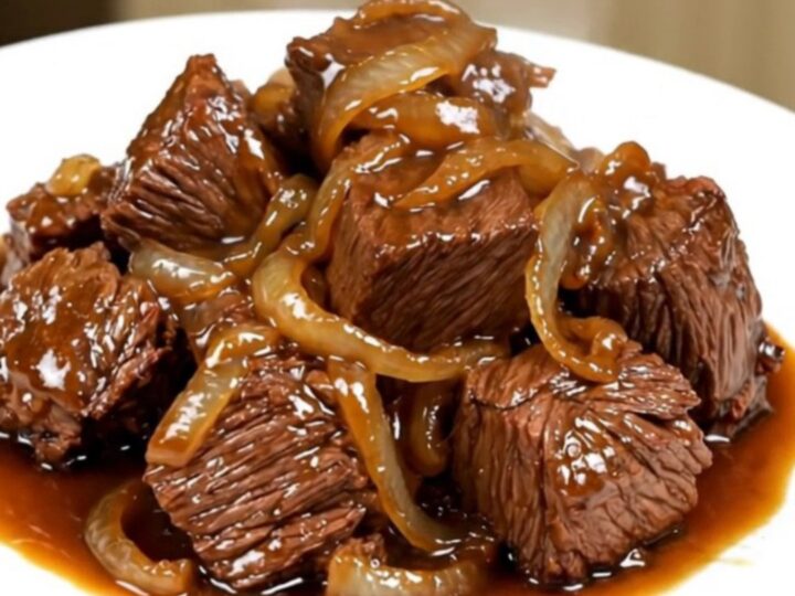 A close-up of tender beef chunks and caramelized onions served on a white plate with a glossy, savory sauce.