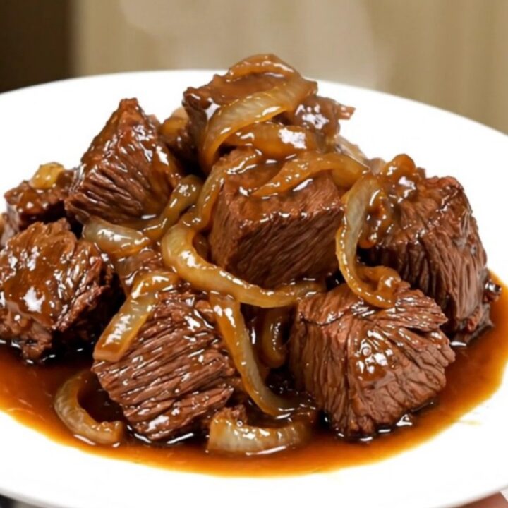 A close-up of tender beef chunks and caramelized onions served on a white plate with a glossy, savory sauce.