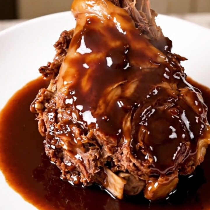 Two tender lamb shanks in a rich, dark red wine sauce with shallots on a white plate.