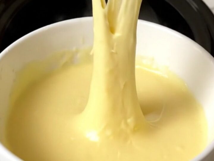 A slow cooker filled with creamy, melted three-cheese fondue on a speckled quartz countertop.