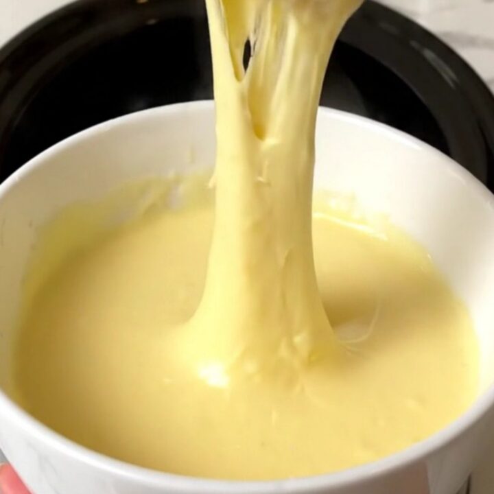 A slow cooker filled with creamy, melted three-cheese fondue on a speckled quartz countertop.