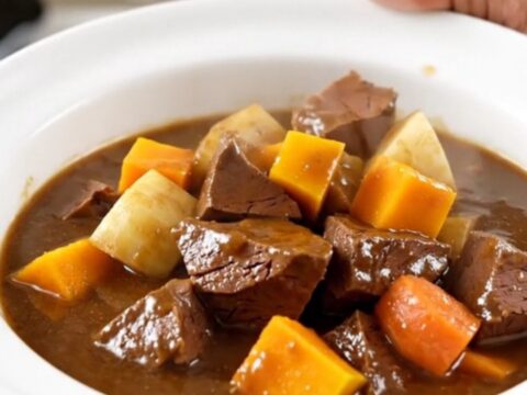 A top-down view of a black oval slow cooker filled with raw veal cubes and colorful root vegetables.