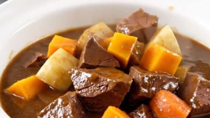 A top-down view of a black oval slow cooker filled with raw veal cubes and colorful root vegetables.