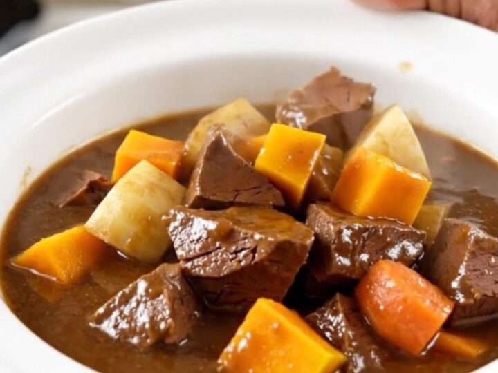 A top-down view of a black oval slow cooker filled with raw veal cubes and colorful root vegetables.