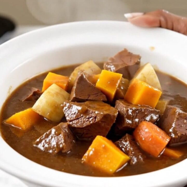 A top-down view of a black oval slow cooker filled with raw veal cubes and colorful root vegetables.