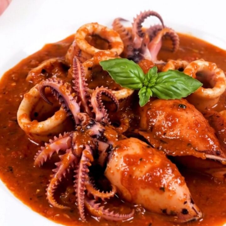 A top-down view of raw cuttlefish, sliced onions, garlic, and tomato sauce in a slow cooker insert.