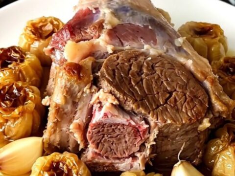 A tender, slow-cooked leg of lamb served on a white platter with soft garlic cloves.