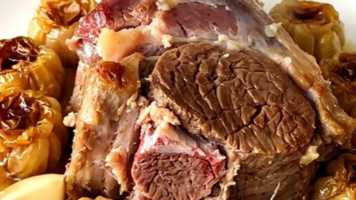 A tender, slow-cooked leg of lamb served on a white platter with soft garlic cloves.