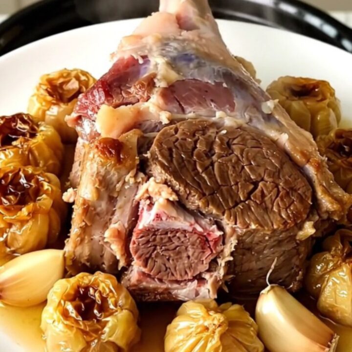 A tender, slow-cooked leg of lamb served on a white platter with soft garlic cloves.