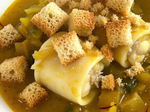 A white bowl filled with monkfish stew, glossy sauce, leeks, and carrots, topped with golden croutons.