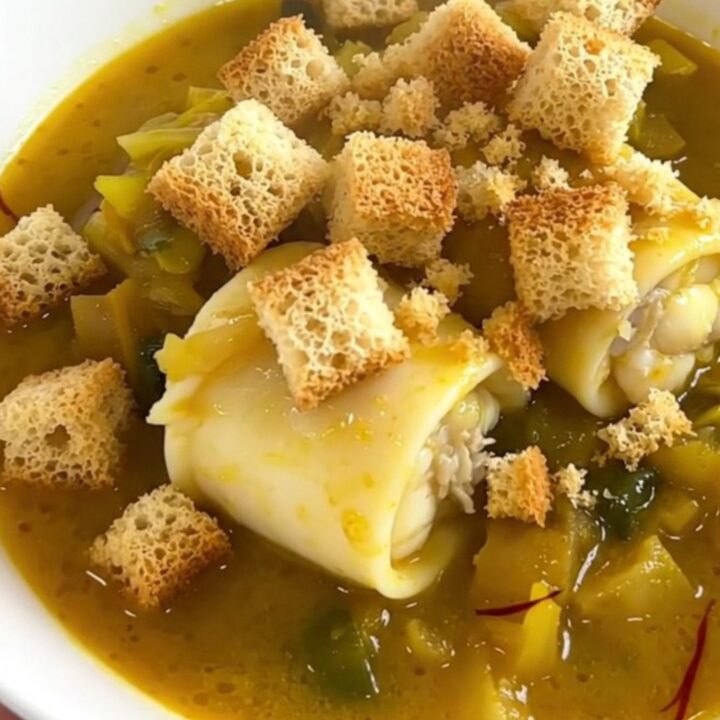 A white bowl filled with monkfish stew, glossy sauce, leeks, and carrots, topped with golden croutons.