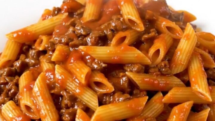 A top-down view of a slow cooker filled with raw ground beef, dry penne pasta, and tomato sauce, ready to cook.