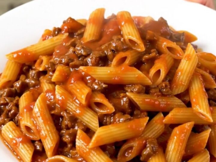 A top-down view of a slow cooker filled with raw ground beef, dry penne pasta, and tomato sauce, ready to cook.