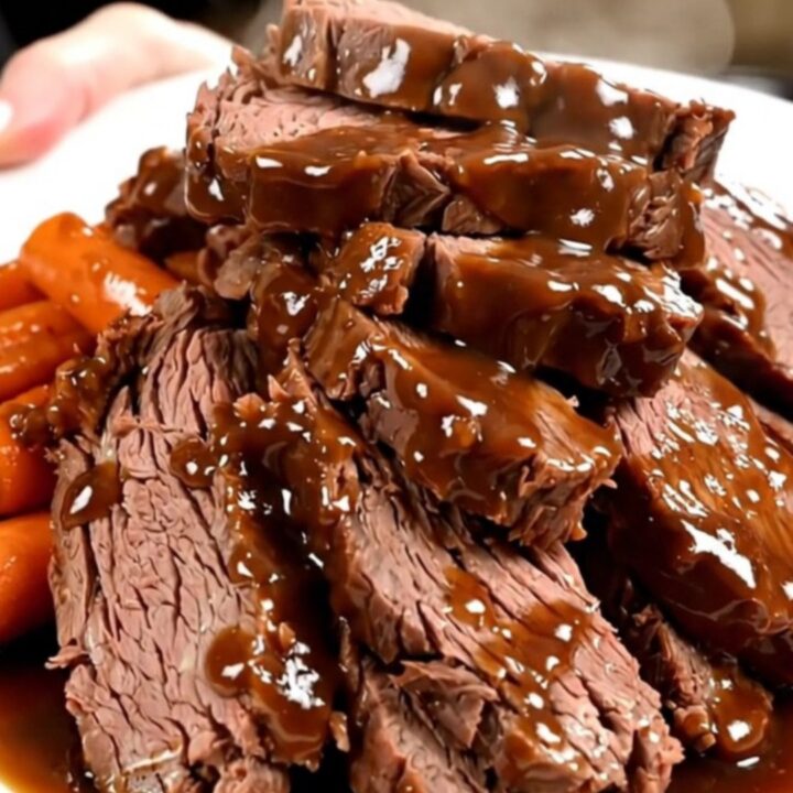 A close-up of tender, sliced beef chuck roast covered in a rich, glossy dark sauce with glazed carrots on a white plate.
