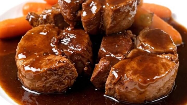 A close-up view of tender braised pork cheeks served with sliced carrots in a rich red wine sauce on a white plate.