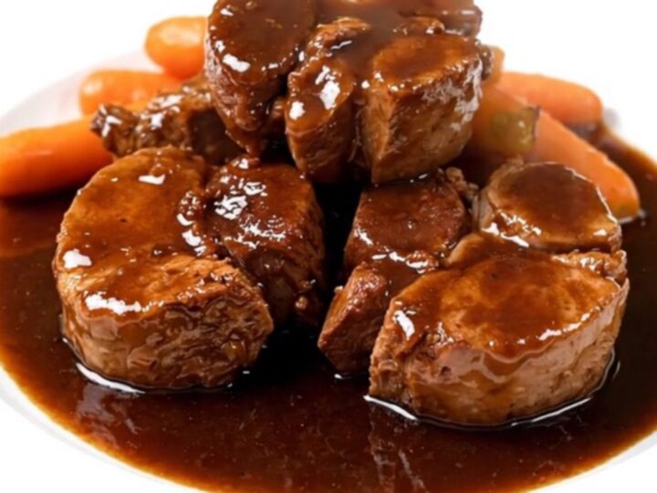 A close-up view of tender braised pork cheeks served with sliced carrots in a rich red wine sauce on a white plate.
