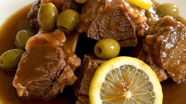 A black slow cooker filled with pieces of cooked lamb, green olives, and lemon slices on a quartz countertop.