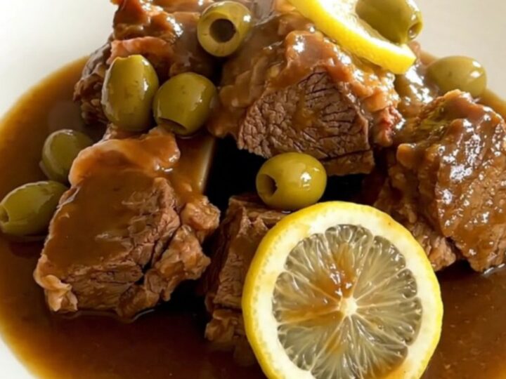 A black slow cooker filled with pieces of cooked lamb, green olives, and lemon slices on a quartz countertop.