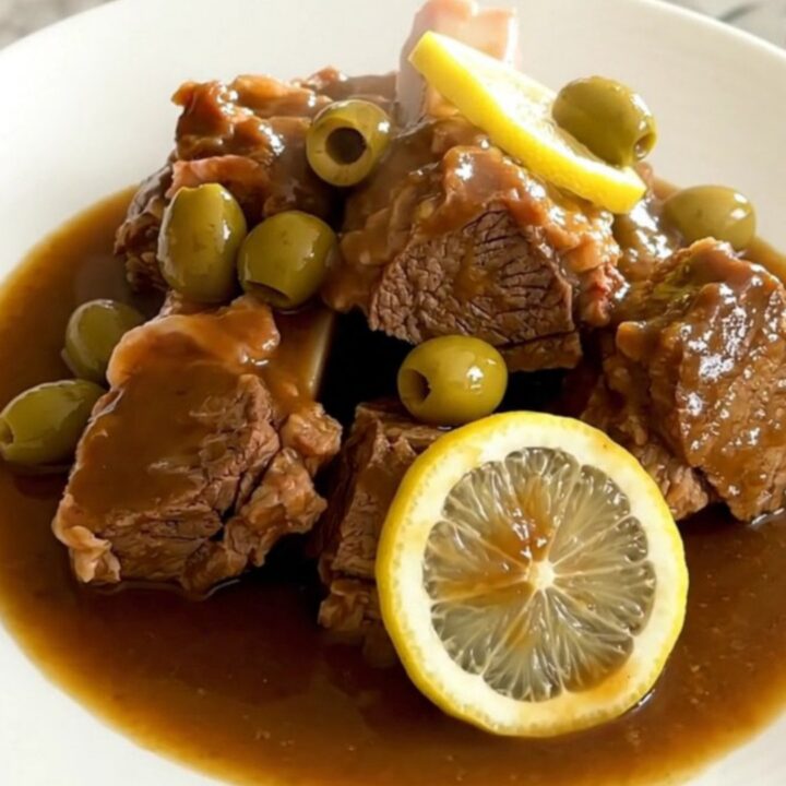 A black slow cooker filled with pieces of cooked lamb, green olives, and lemon slices on a quartz countertop.