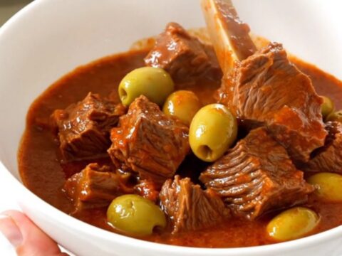 A top-down view of a steaming beef stew with olives in a slow cooker on a white countertop.