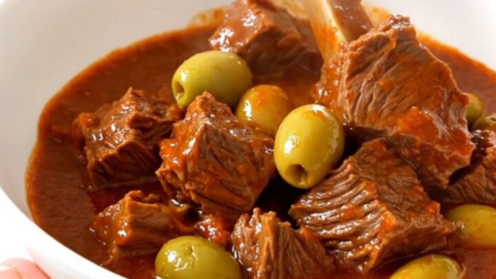 A top-down view of a steaming beef stew with olives in a slow cooker on a white countertop.