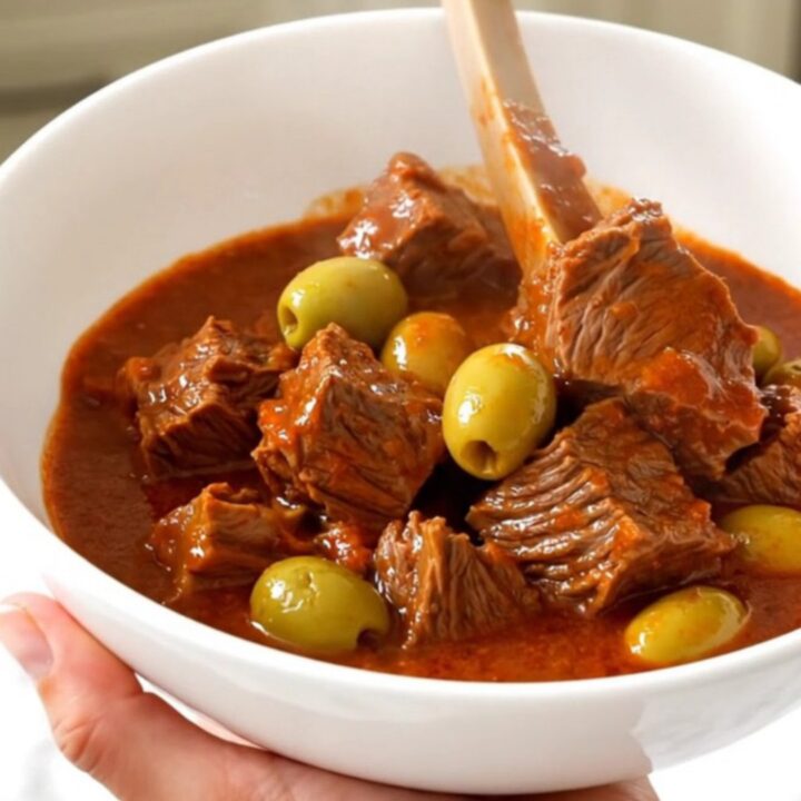 A top-down view of a steaming beef stew with olives in a slow cooker on a white countertop.