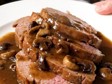 A close-up of a tender, sliced veal roast served with wild mushrooms and a rich, glossy gravy on a white plate.