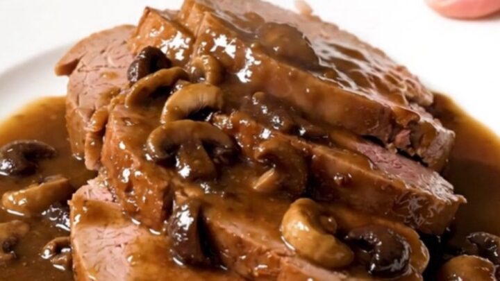A close-up of a tender, sliced veal roast served with wild mushrooms and a rich, glossy gravy on a white plate.