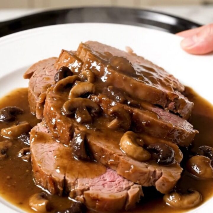 A close-up of a tender, sliced veal roast served with wild mushrooms and a rich, glossy gravy on a white plate.