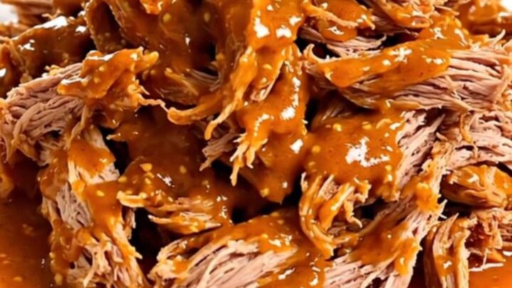 A close-up of tender, shredded pork shoulder coated in a glossy, spicy red mustard-tomato sauce on a white plate.