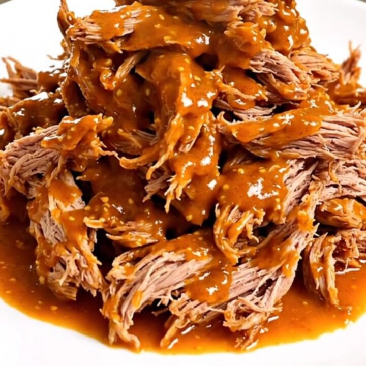 A close-up of tender, shredded pork shoulder coated in a glossy, spicy red mustard-tomato sauce on a white plate.