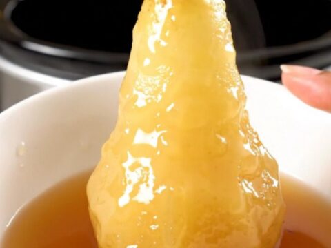 Four whole peeled pears simmering in a dark cider syrup inside a slow cooker on a white countertop.