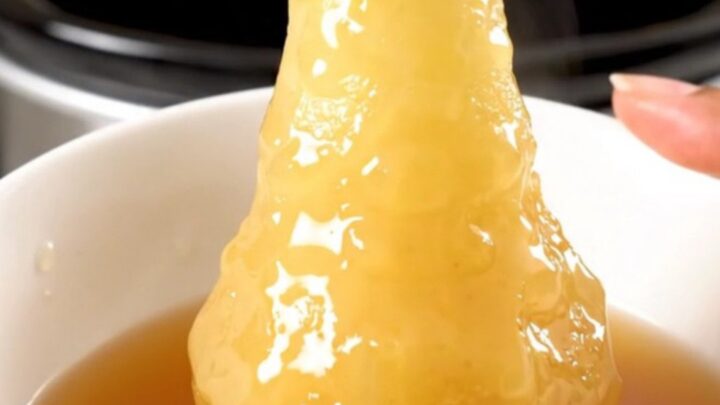 Four whole peeled pears simmering in a dark cider syrup inside a slow cooker on a white countertop.