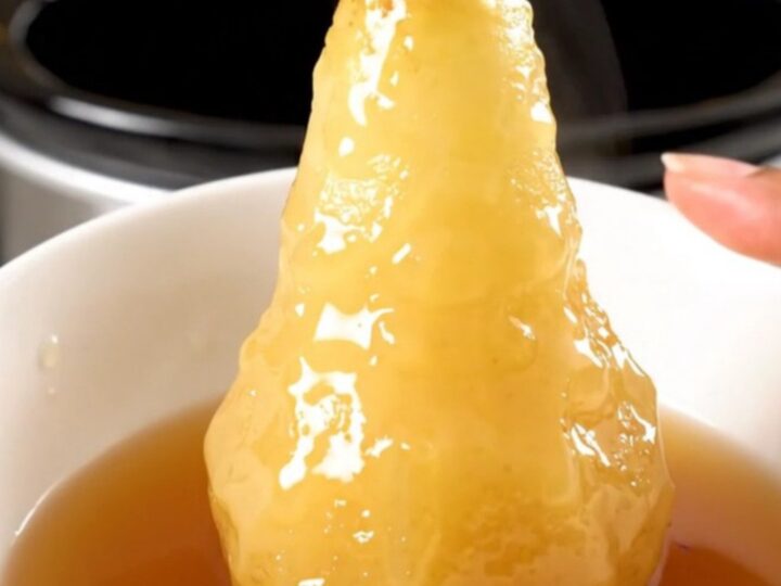 Four whole peeled pears simmering in a dark cider syrup inside a slow cooker on a white countertop.