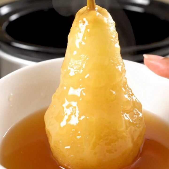 Four whole peeled pears simmering in a dark cider syrup inside a slow cooker on a white countertop.