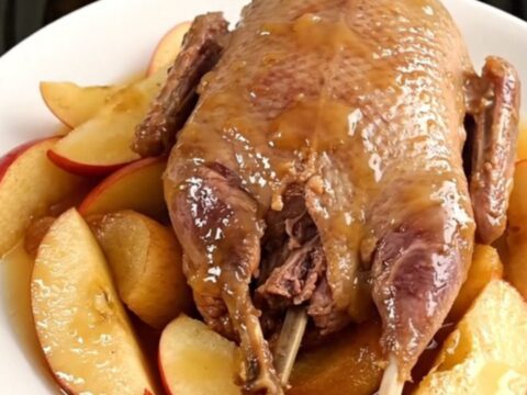 A rustic plate featuring slow-cooked guinea fowl pieces, tender apple wedges, and glazed shallots in a savory sauce.