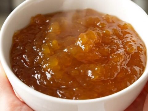 A rustic bowl of glossy, golden-orange pumpkin jam sitting on a white speckled countertop with soft kitchen lighting.