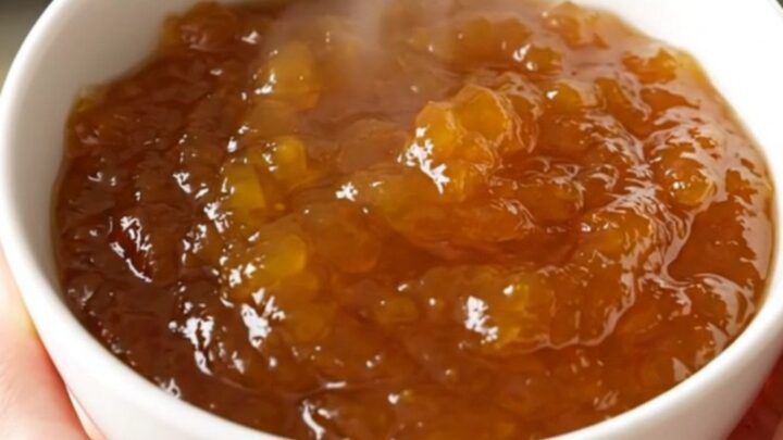 A rustic bowl of glossy, golden-orange pumpkin jam sitting on a white speckled countertop with soft kitchen lighting.