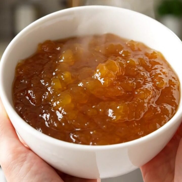 A rustic bowl of glossy, golden-orange pumpkin jam sitting on a white speckled countertop with soft kitchen lighting.