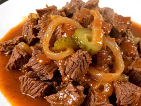 A close-up of a steaming, glossy beef miroton served on a white plate with onion and pickle slices visible.