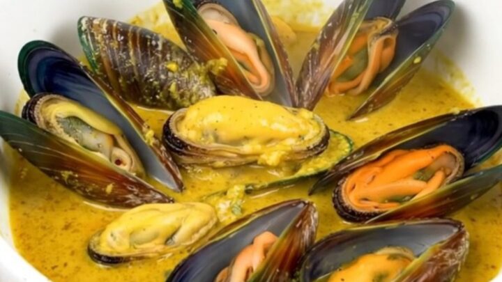 A white bowl filled with steamed mussels in a creamy, golden curry sauce, set against a blurred kitchen background.