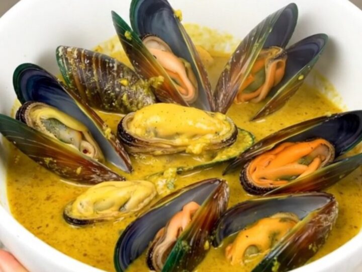 A white bowl filled with steamed mussels in a creamy, golden curry sauce, set against a blurred kitchen background.