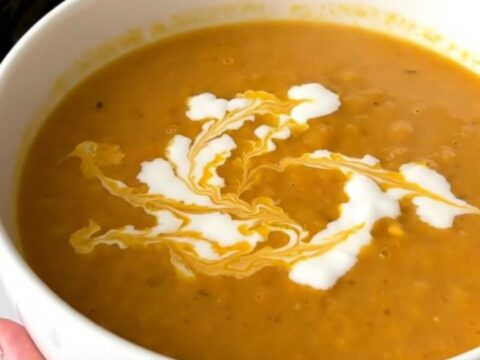 A white ceramic bowl filled with vibrant orange, creamy red lentil and carrot soup, garnished with a swirl of coconut milk.