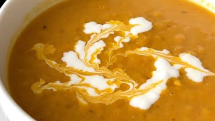 A white ceramic bowl filled with vibrant orange, creamy red lentil and carrot soup, garnished with a swirl of coconut milk.