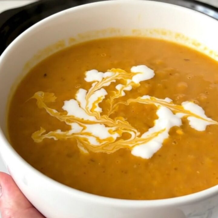 A white ceramic bowl filled with vibrant orange, creamy red lentil and carrot soup, garnished with a swirl of coconut milk.