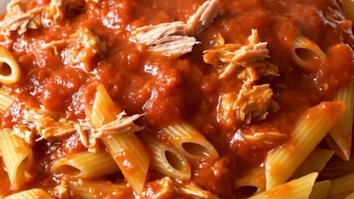 A steaming bowl of penne pasta tossed in a rich, glossy red tomato sauce with visible flakes of tuna.