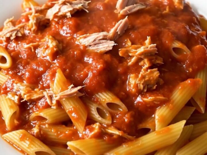 A steaming bowl of penne pasta tossed in a rich, glossy red tomato sauce with visible flakes of tuna.