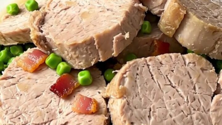 A close-up of sliced pork tenderloin medallions topped with green peas and bacon lardons on a white plate.